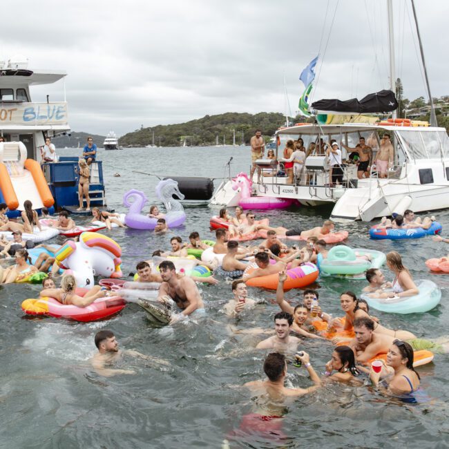 A lively scene of people enjoying a party in the water. They are surrounded by various colorful inflatable floats, including unicorns and animals. Two boats named "Barefoot" and "Blue" are anchored nearby. The sky is overcast.