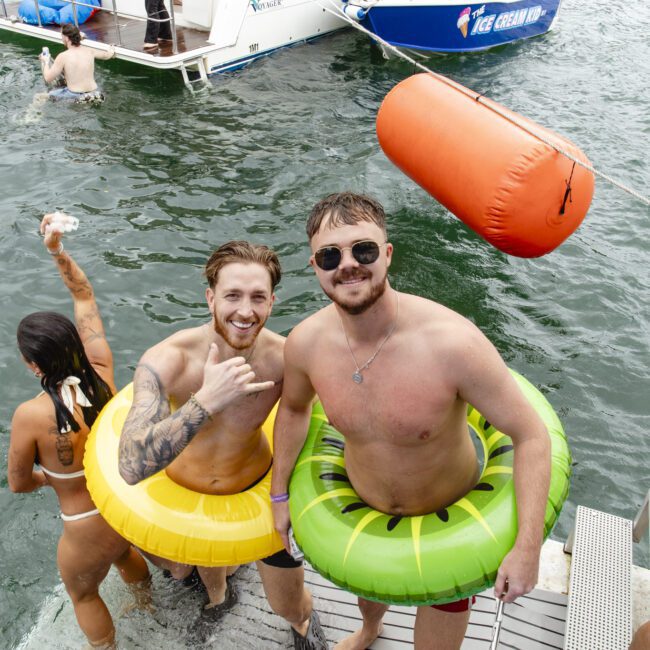 Two men wearing inflatable rings are standing on boat steps leading into a lake. One wears a yellow ring, the other a green one. They're smiling at the camera. Nearby, others enjoy the water and a boat is in the background.