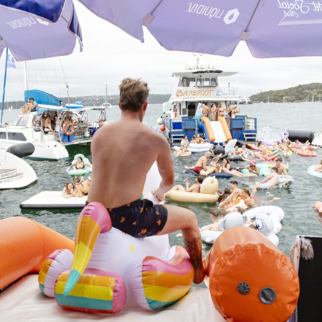 A person sits on an inflatable unicorn float, surrounded by other pool floats and people in the water. In the background, a boat party is happening with many participants and inflatables. Large umbrellas provide shade from above.