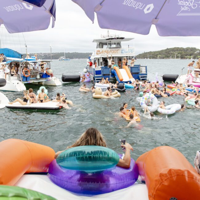 A group of people enjoying a pool party on the water, floating on inflatables. A boat is anchored nearby with the sign "BAREFOOT & LIVE." Several people are holding drinks, and there are branded umbrellas overhead.