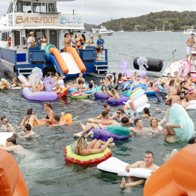 A large group of people enjoying a party on a boat and in the water, surrounded by colorful inflatable floats. The boat, named Barefoot Blue, is docked in a scenic area with trees and other boats in the background.