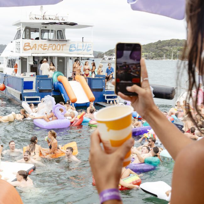 A lively boat party scene with people swimming and lounging on inflatable floats in the water. A large boat with "Barefoot Blue" on the side is anchored nearby. A person holds a phone and cup in the foreground, capturing the festive atmosphere.