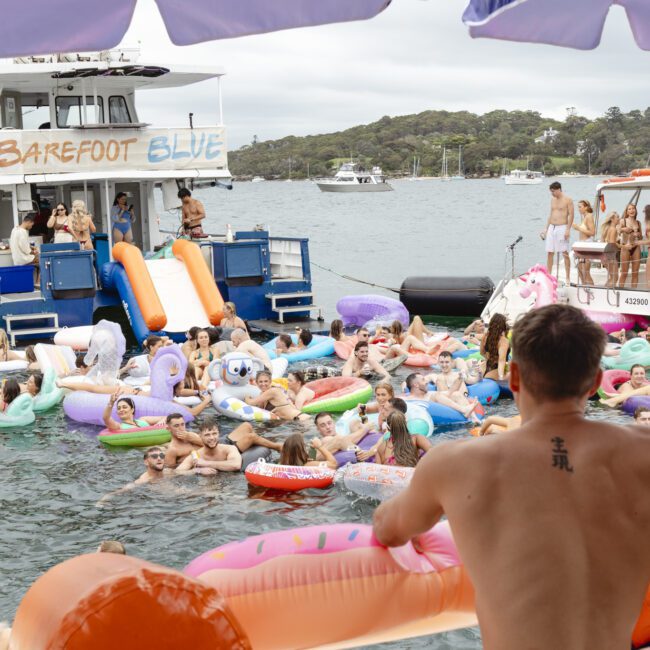 A crowded scene on a lake with people on colorful inflatable floats. A man sits on a float at the bottom right, facing a busy ferry named "Barefoot Blue." Overcast sky and wooded shoreline in the background.