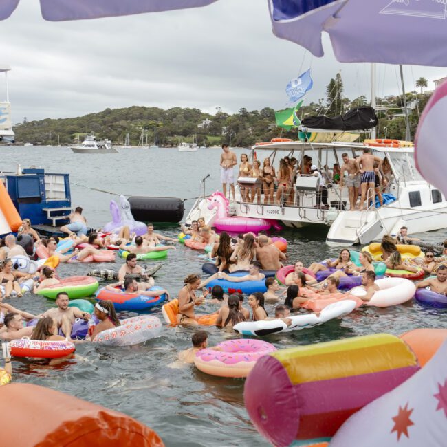 A crowded pool party scene with numerous people enjoying the water on inflatable pool floats shaped like unicorns, donuts, and other objects. Several boats are anchored nearby, and the sky is cloudy. Umbrellas and trees are in the background.