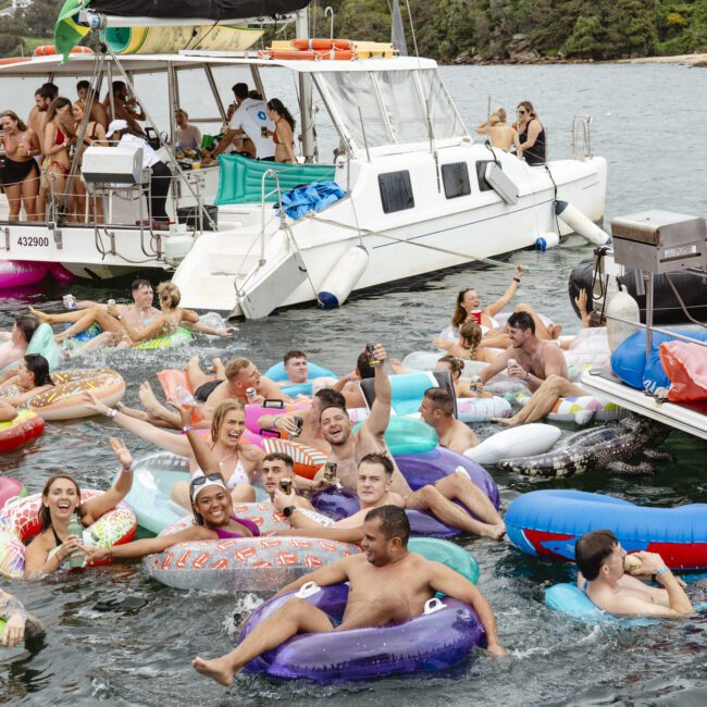 A lively group of people enjoying a floating party in the water. They're on colorful inflatable toys, including a unicorn and various pool floats, near two boats. The setting is a sunny day with trees visible in the background.