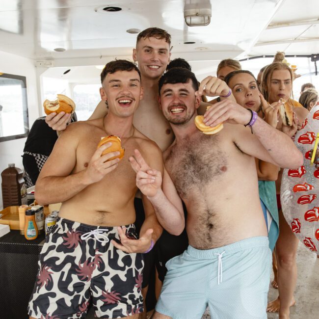 A group of friends on a boat, smiling and holding burgers. Two men in swim trunks pose at the front, with others in the background. The atmosphere is cheerful and relaxed. Sunlight filters in from outside.