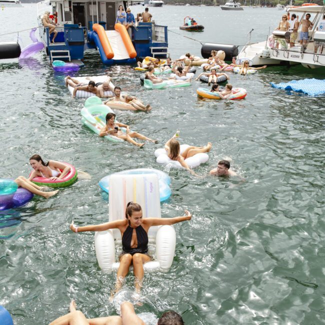A group of people relaxing on inflatable tubes in the water near a boat named "Barefoot Blue." The scene is lively, with people enjoying a sunny day, and the boat has slides extending into the water. Hills and trees are visible in the background.