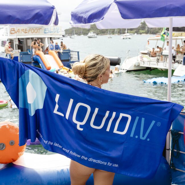 A person draped in a Liquid I.V. towel stands by the water. Several people are seen on nearby boats and inflatables, with a background of calm water and a partly cloudy sky. Umbrellas and pool floats add a festive vibe.