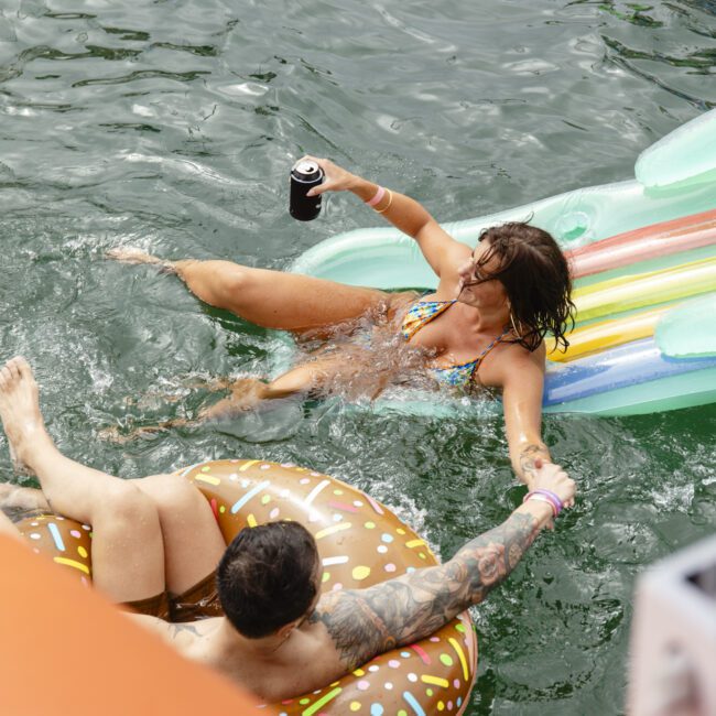 A woman in a patterned bikini is lounging on a striped inflatable raft in a pool, holding a drink. Beside her, a person with tattoos is floating in a donut-shaped tube, extending a hand to her.