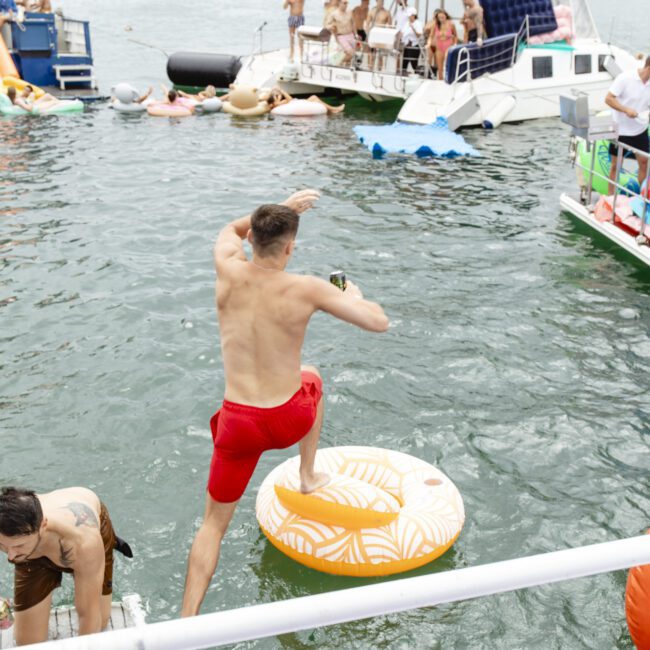 A man in red shorts jumps onto an inflatable orange float on a lake, surrounded by boats and several people. The scene is lively with multiple people enjoying the water and watching.