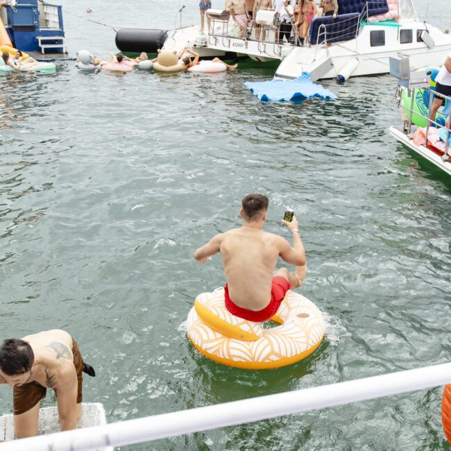 A man in red shorts sits on an inflatable float with a phone in hand, surrounded by people on boats and floats in a lake. Various inflatables, including doughnuts and a pizza slice, are in the water. A lively gathering is underway.