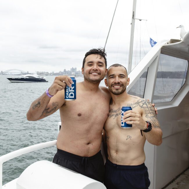 Two men in swim trunks stand side by side on a boat. Both are smiling and holding drinks. They have tattoos and appear relaxed. The ocean and part of a city skyline are visible in the background under a cloudy sky.