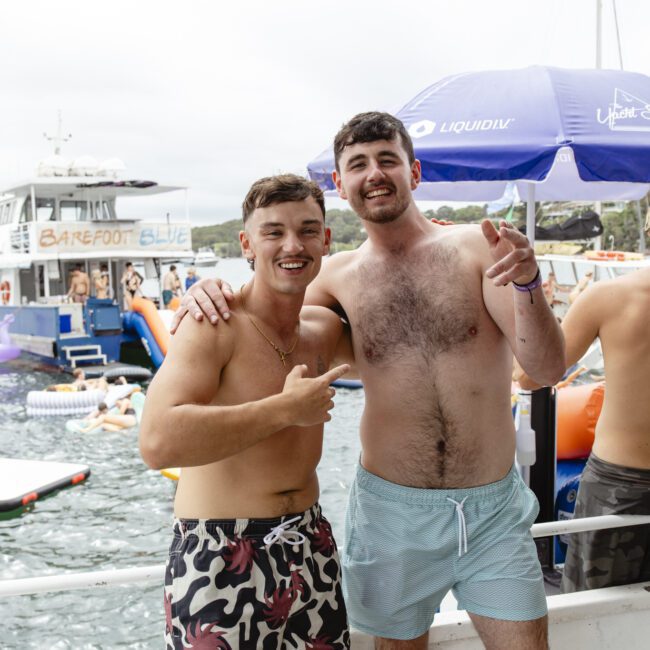 Two men smiling and posing on a boat near the water. One wears patterned swim trunks and the other light blue shorts. In the background, there's another boat with people, inflatables, and umbrellas. It's a lively, sunny scene.