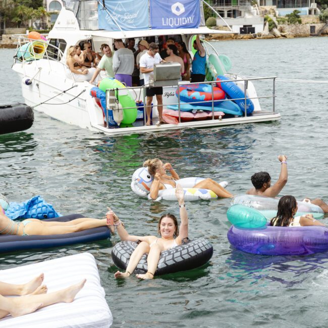 People are enjoying a sunny day on the water near a boat. Some are floating on inflatables while others socialize on the vessel. The backdrop features houses and trees on a hillside.