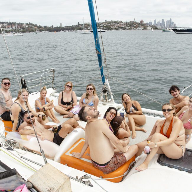 A group of people relaxing and smiling on a sailboat deck, wearing swimsuits. They are gathered around the boat's edge, with a scenic harbor and distant city skyline in the background. The atmosphere is casual and cheerful.
