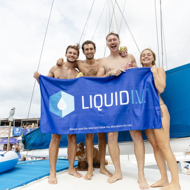 Four people in swimwear stand on a boat holding a blue "Liquid I.V." banner. They appear happy and are surrounded by a festive atmosphere. The background shows buildings and another boat on the water under a cloudy sky.