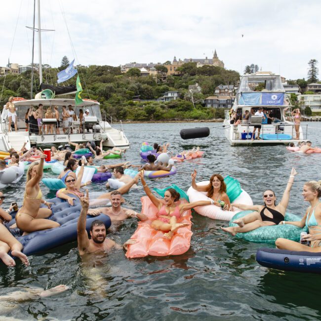 A group of people enjoying a sunny day on a lake, floating on various inflatable rafts shaped like sofas, a unicorn, and a watermelon slice. Boats are anchored nearby, and houses and trees are visible in the background.