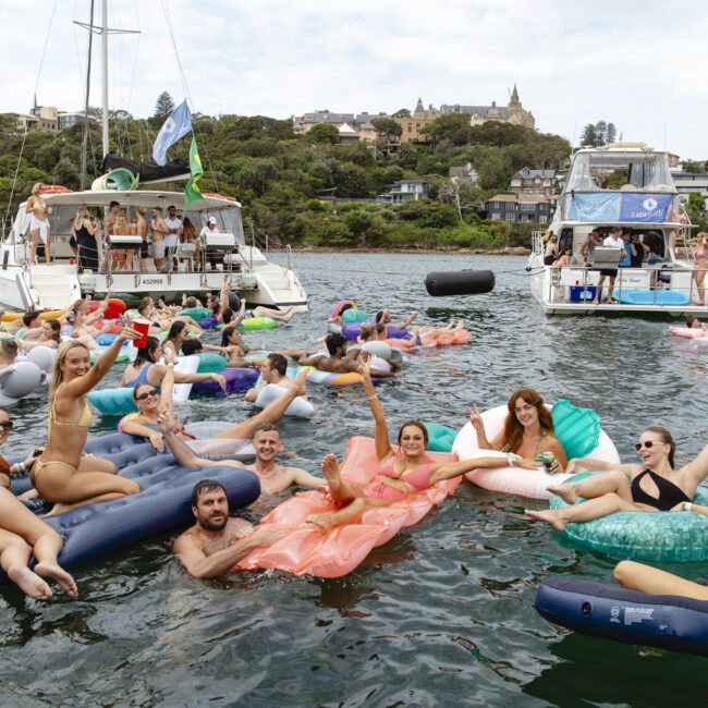 A group of people relax on inflatable loungers in the water near boats. They're enjoying a sunny day, smiling and holding drinks. The shore with buildings and trees is visible in the background.