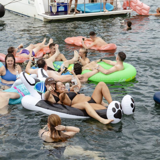 A group of people enjoying a sunny day on a lake, floating on colorful inflatable rafts, including a panda and other animal shapes. They are relaxed, laughing, and socializing near a boat.
