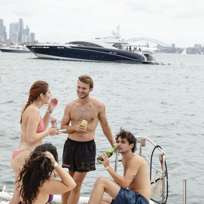 Four people in swimsuits enjoy drinks and laughter on a boat. The water is calm, with a large yacht and a city skyline, including a bridge, in the background. The scene is relaxed and cheerful.