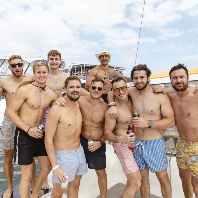 A group of ten shirtless men in swim trunks are posing on a boat, smiling and holding drinks. The sky is partly cloudy, and there are other boats in the background.