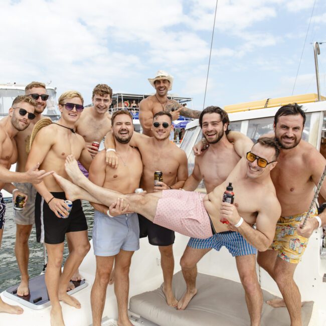 A group of ten men on a boat, smiling and posing. Some wear sunglasses, swim trunks, and hats. The group playfully holds one person horizontally. There are other boats and a cloudy sky in the background.