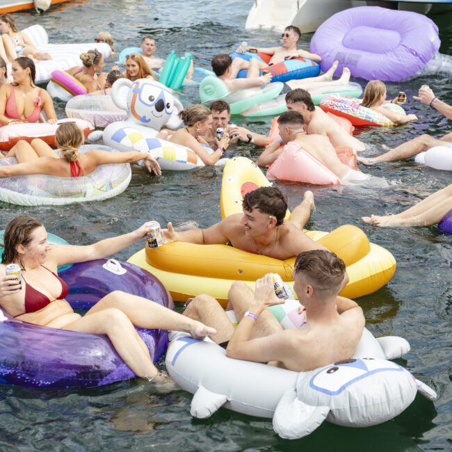 A lively group of people enjoying time in the water using various inflatable pool floats. Many hold beverages and smile, with colorful inflatables shaped like animals and food surrounding them. The atmosphere is festive and relaxed.