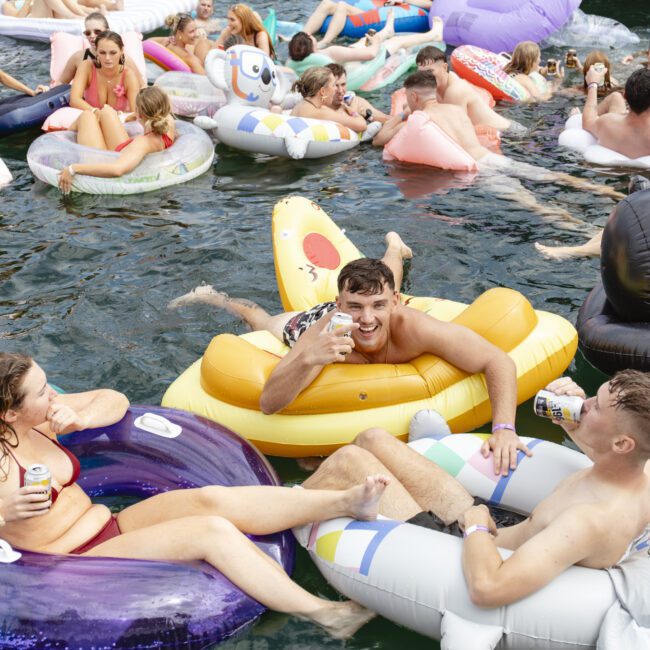 A large group of people enjoying a pool party, lounging on colorful inflatable rafts. They are smiling, holding drinks, and wearing swimwear. The water is crowded with floats of various shapes and colors, creating a lively atmosphere.