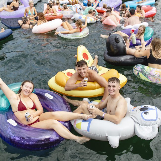 A group of people having fun on water floats in a lake. They are wearing swimwear and holding drinks, smiling and waving. The scene is lively with a variety of colorful inflatable rafts, including a pizza and animal shapes.
