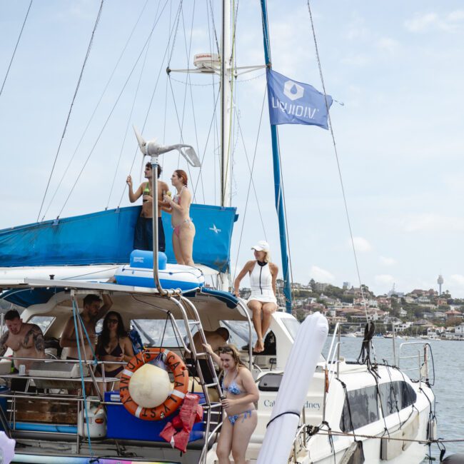 A group of people enjoying a day on a sailboat adorned with inflatable toys. Several individuals are on the deck, some in swimwear, under a blue flag. The backdrop features a coastal town with buildings and greenery. The sky is partly cloudy.