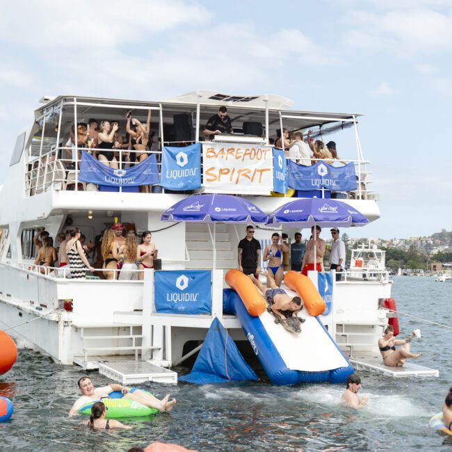 A lively boat party with people enjoying music and drinks on a two-story vessel. Several people are sliding into the water from inflatable slides, while others float nearby on inflatable structures. The sky is partly cloudy.