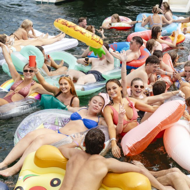 A lively group of people enjoying a sunny day on a lake, floating on inflatable rafts shaped like pizza and animals. They are smiling, socializing, and holding drinks, with a pontoon boat visible in the background. The atmosphere is fun and festive.