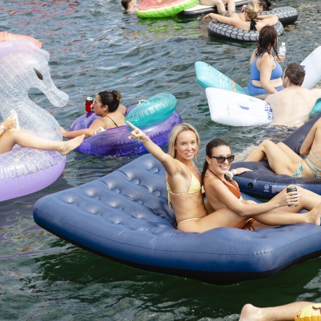 People relaxing on colorful inflatable rafts in water near a boat. A person in a yellow bikini sits on a blue raft, smiling and gesturing toward another person wearing sunglasses. Others enjoy various inflatable shapes like a unicorn and watermelon slice.
