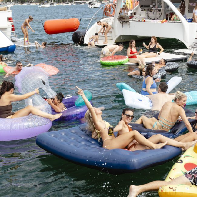 People enjoying a sunny day on inflatable floats in the water around docked boats. Some are swimming, while others relax on colorful rafts, surrounded by boats and a distant shoreline with buildings and trees.