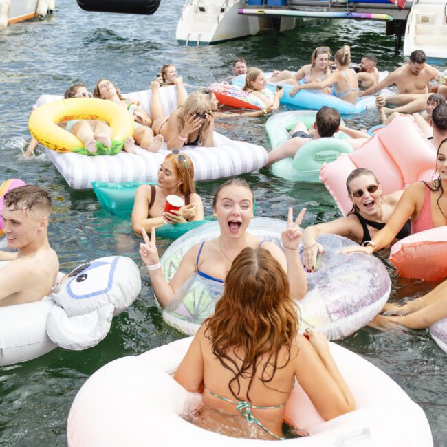 A group of people in swimsuits are enjoying a sunny day in the water, lounging on inflatable rings and floats. Boats are visible in the background. Everyone is smiling and relaxed, with one person playfully flashing a peace sign.