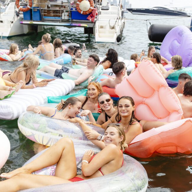 A group of people relaxing on colorful inflatable rafts in a sunny, busy harbor. They're smiling and chatting on the water with sailboats and a city skyline in the background.