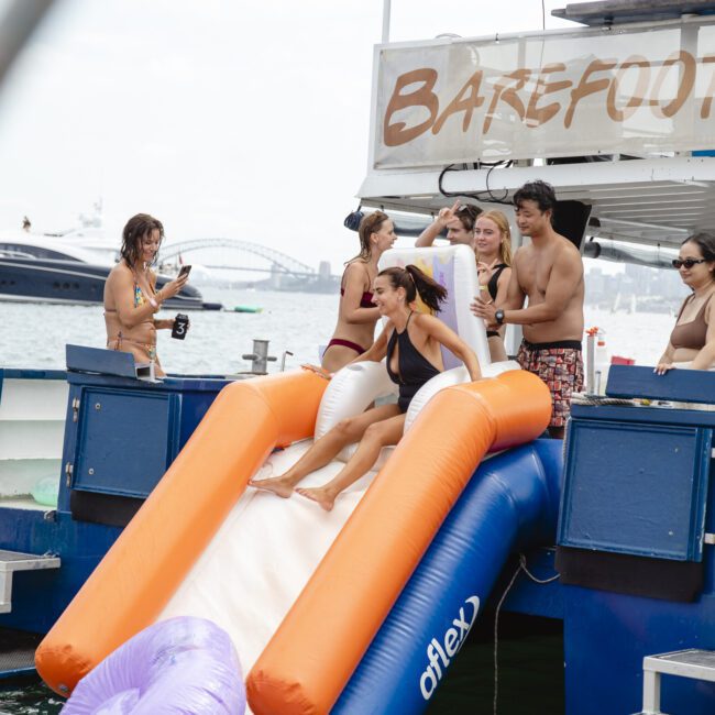 A group of people enjoying a yacht party. A woman is sliding down a large inflatable orange slide into the water. Others are standing nearby, chatting and watching. Bridge visible in the background. Party atmosphere with bright outdoor setting.