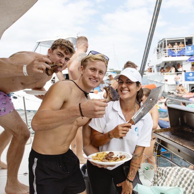 Three people on a boat enjoying grilled meat. Two are holding plates with food, smiling, while a third person stands in the background holding food. Boats and people in the water are seen in the background. It's a lively, sunny scene.