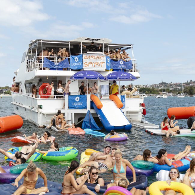 A group of people enjoying a sunny day on the water. They are floating on colorful inflatable rafts near a large boat, which has people on its deck. The boat has a sign reading "Barefoot Spirit." The sky is partly cloudy.