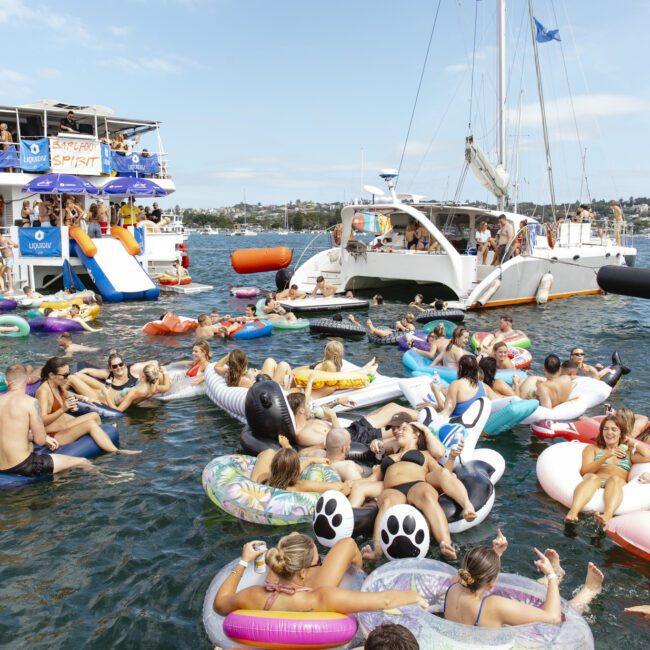 A lively scene on a sunny day with people on inflatable floats in the water, surrounded by boats. Participants are enjoying the gathering and music, with some seated on colorful floats shaped like animals. The atmosphere is festive and relaxed.