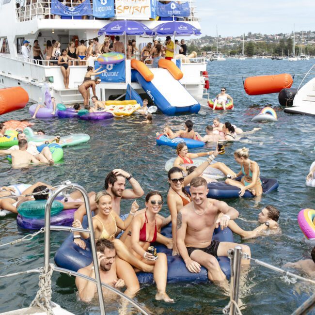 A lively outdoor party scene on a boat and water. People are on inflatable floats, some in swimwear, enjoying sunny weather. A boat, decorated with "Barefoot Spirit" and sponsor banners, is in the background.