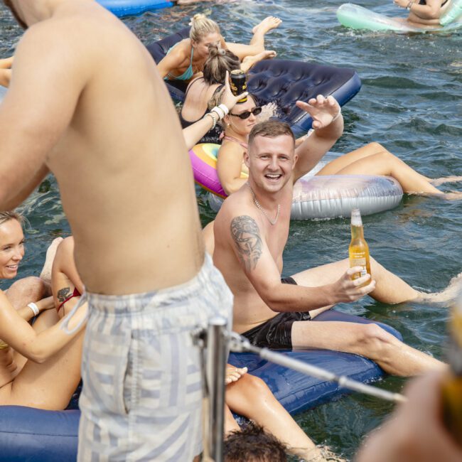 A lively group enjoying a sunny day on the water, lounging on inflatables. People are in swimwear; one man waves and holds a bottle. Various colorful inflatables and laughter fill the scene. The atmosphere is festive and relaxed.