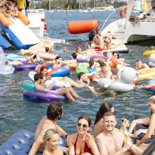 A group of people are floating on colorful inflatables in a sunny lake, surrounded by boats. Some are swimming, while others relax on inflatables and boats. Smiles and laughter are abundant, with a clear blue sky in the background.