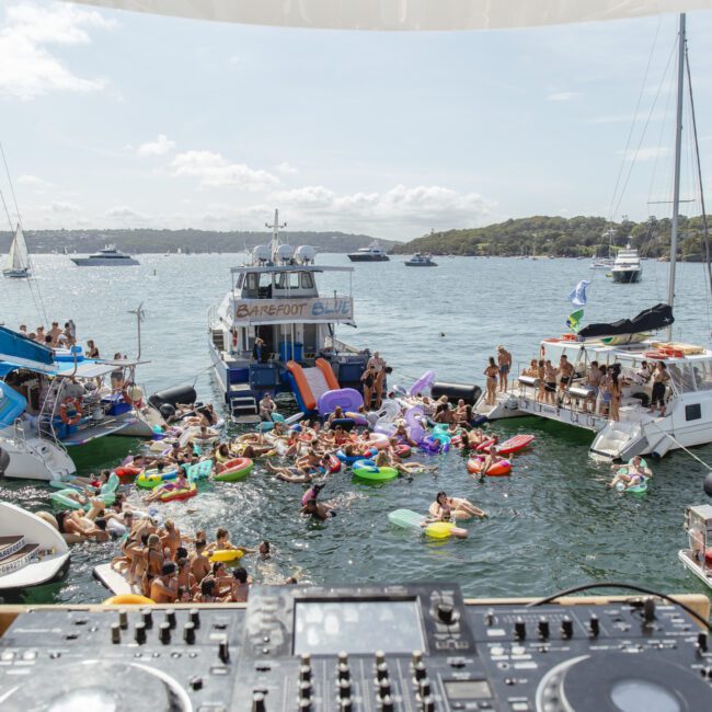View from behind a DJ booth showing a lively boat party. People are on boats and colorful inflatables in the water, with a bright sky and more boats in the background. Reflections of partygoers are visible on a mirrored surface above.
