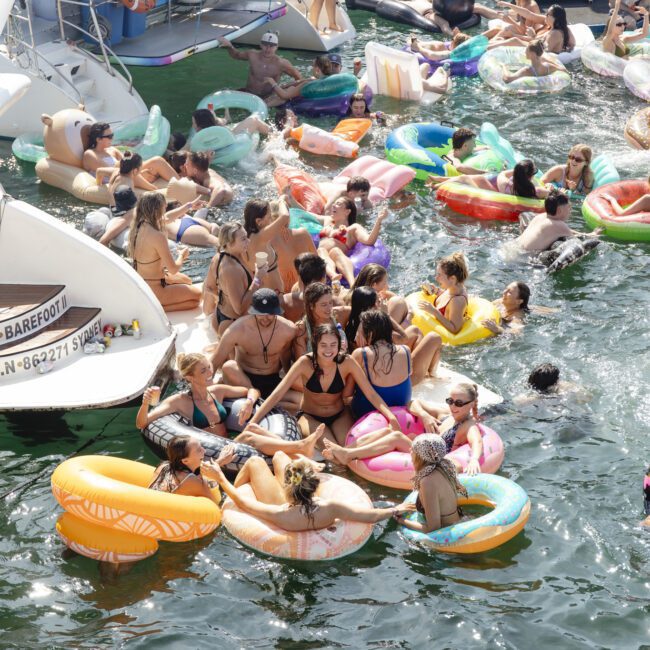 A lively group of people enjoying a sunny day on a body of water, floating on colorful inflatable tubes. Boats are nearby, and everyone is wearing swimwear, creating a festive, summery atmosphere.