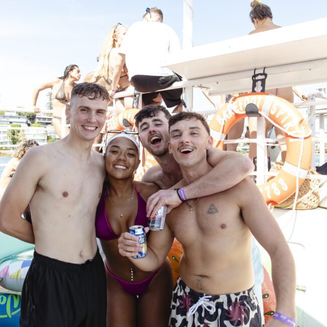 A group of four young adults, three men and one woman, pose and smile on a boat. They are wearing summer attire, and the woman holds a drink. Other people are in the background enjoying the sunny day. Inflatable rings and life preservers are visible.