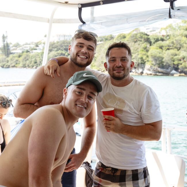 Three men on a boat smile at the camera. They are casually dressed, with one holding a red cup. The background shows water and a tree-lined shore. It's a sunny day.