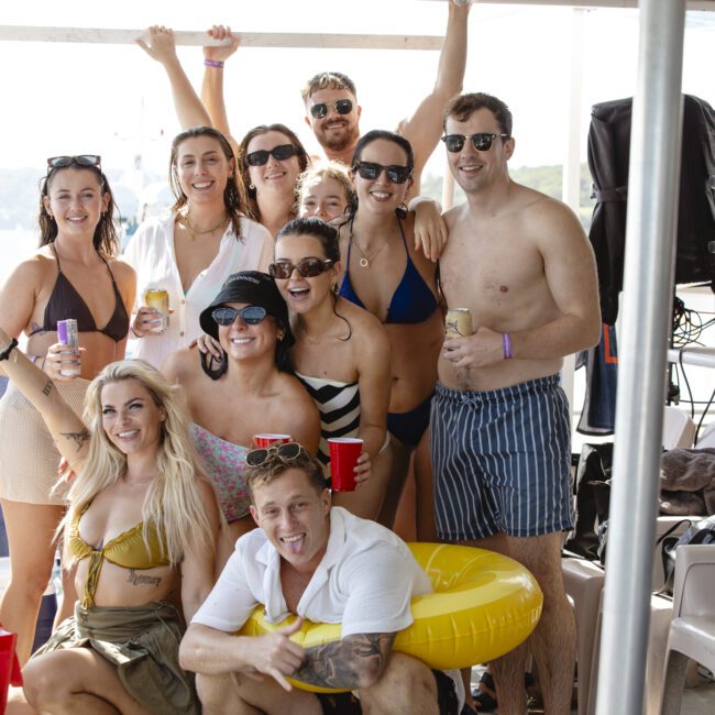 A group of people in swimsuits and sunglasses are smiling and posing on a boat with lakeside scenery. Some are holding drinks, and one person has a yellow float tube around their waist. The atmosphere is relaxed and joyful.