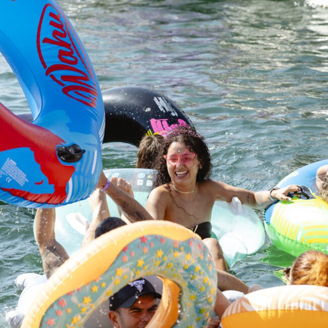 People are joyfully floating on colorful inflatable tubes in a sunlit body of water. One person in pink sunglasses stands out, smiling widely. The atmosphere is lively and fun, with splashes and bright inflatables adding to the scene.