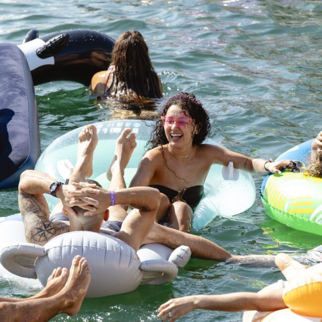 A woman with curly hair and sunglasses joyfully leans over a man on an inflatable float in a lake. Other people are seen on various inflatables nearby, enjoying the water and sunshine.